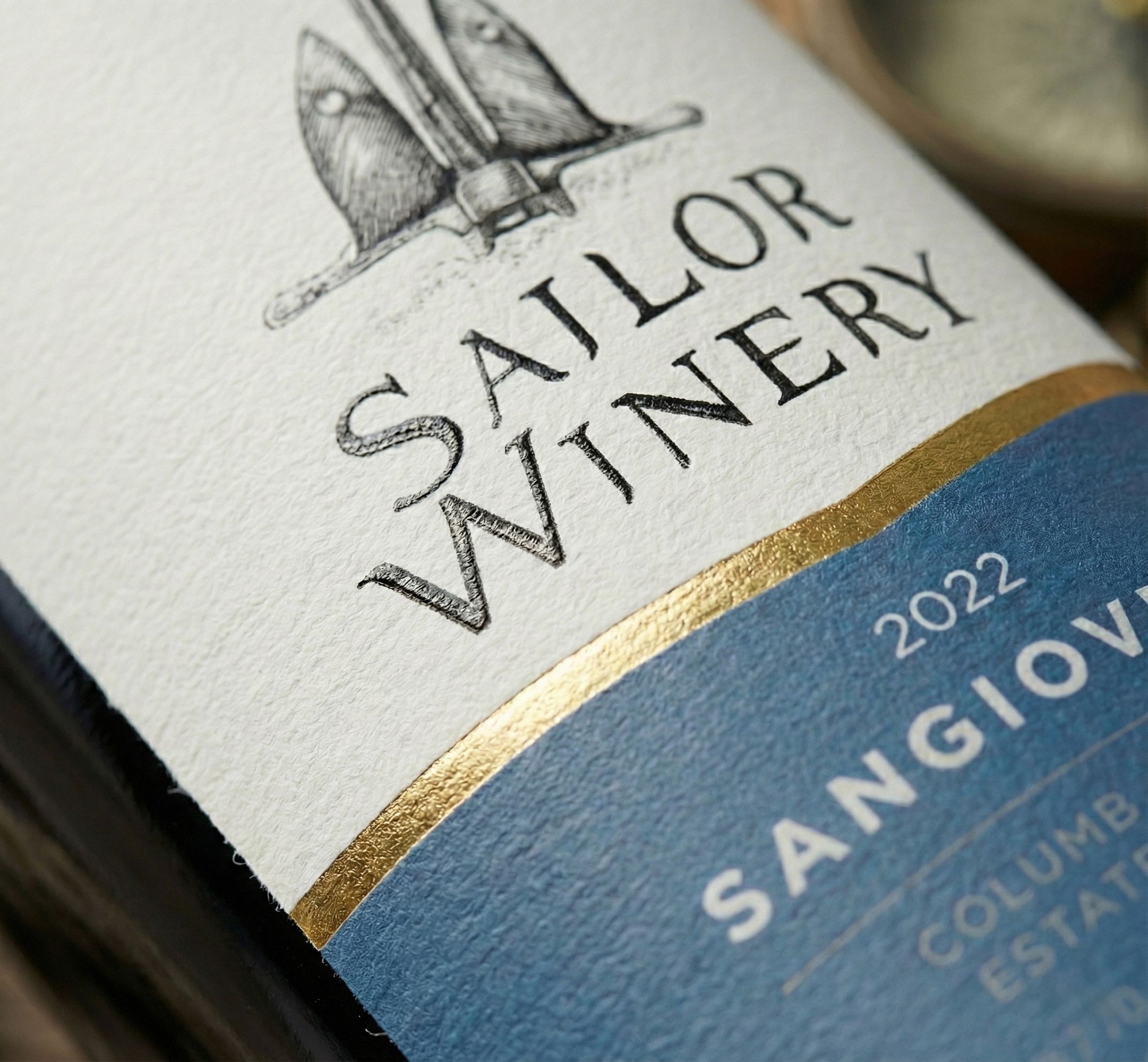 The Sangiovese label in close-up: drawn lettering pressed into uncoated stock, gold wave running into the blue panel