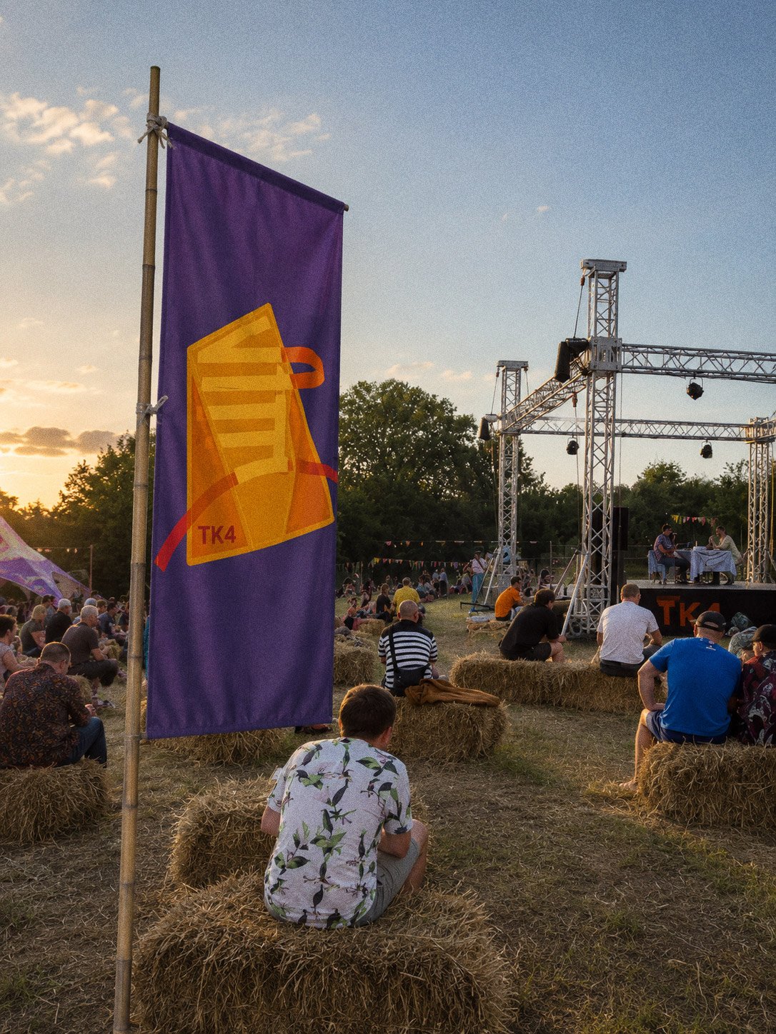 A purple TK4 flag carrying the abstract mark, people sitting on hay bales in front of the stage as the sun goes down