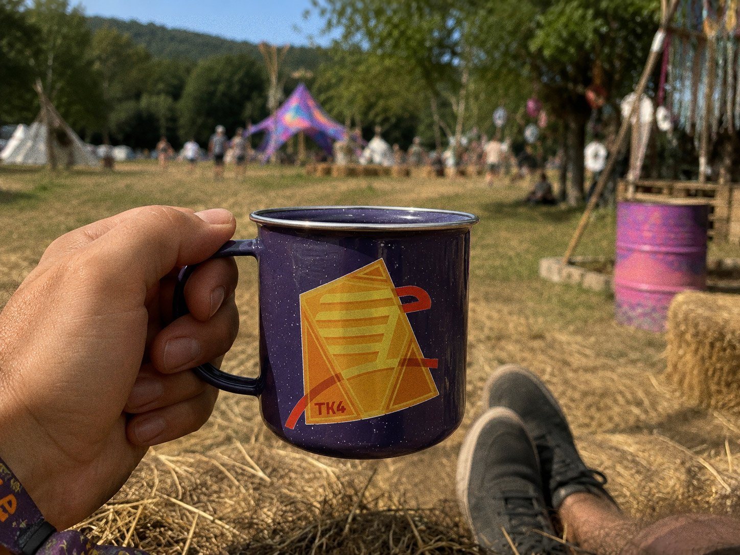 A purple enamel camp mug held up in hand, the abstract TK4 mark on its side, tipis and painted barrels blurred behind